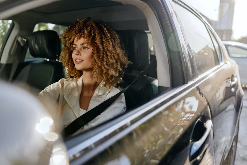 Curly woman driving car safely with seatbelt fastened, focused on road, wearing light blouse under natural sunlight with reflections on car exterior glass.