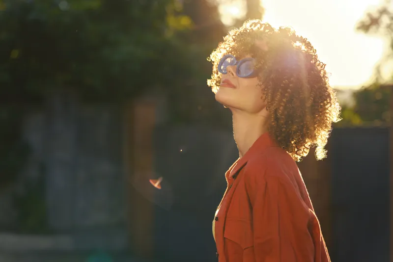 Woman lifestyle portrait outdoors in sunlight wearing orange jacket and sunglasses, relaxed confident mood, backlit glow and soft breeze