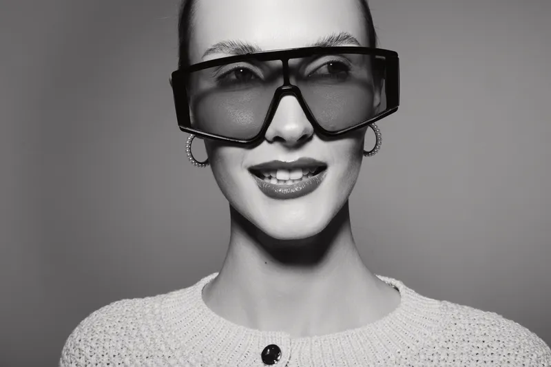 Portrait of a young white woman with pale skin wearing oversized sunglasses and earrings, smiling slightly on grey background.