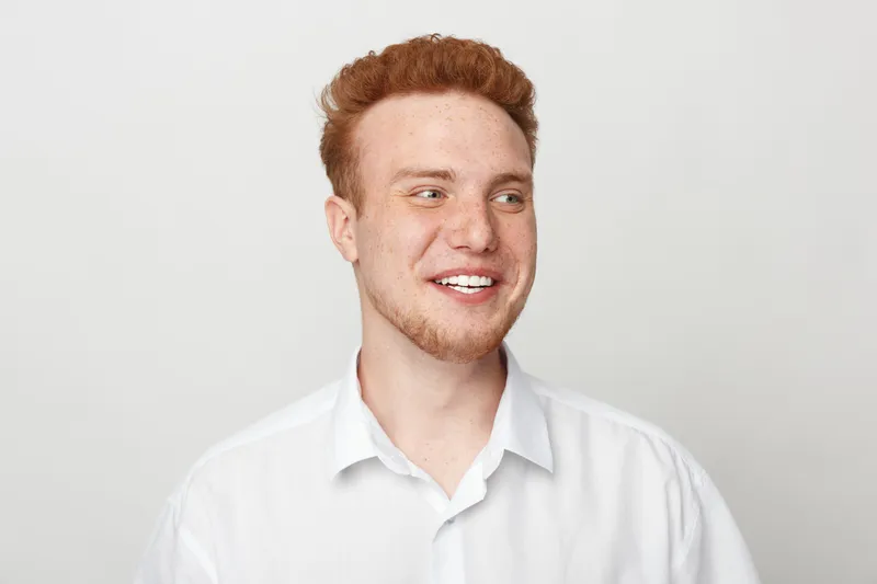 Smiling young man with red hair and beard wearing white shirt looks away on white background. Portrait of happy male model with casual style and positive expression in studio lighting.