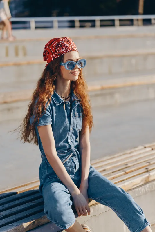 Lifestyle woman in modern boho-western denim street style wearing blue sunglasses and red bandana, sitting outdoors enjoying sunny day with film color warmth and casual vibe.