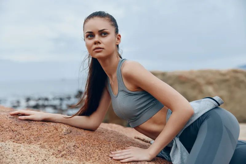Athletic young woman in sportswear, posing on rocky beach, focused expression showcasing determination and strength, ideal for fitness and wellness themes
