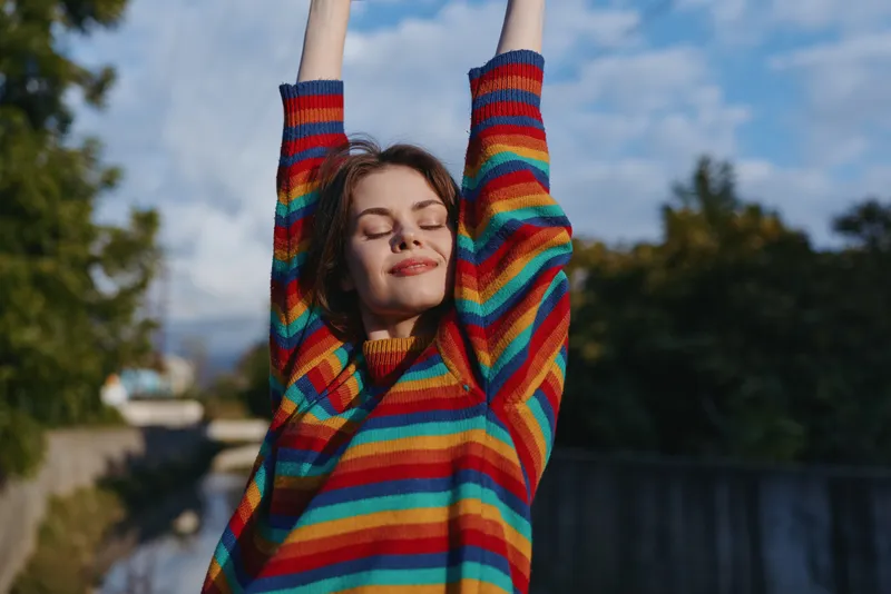 Woman in a rainbow sweater stretching outdoors with eyes closed, smiling portrait of relaxation and joy, casual fashion, sunny day and peaceful urban nature background
