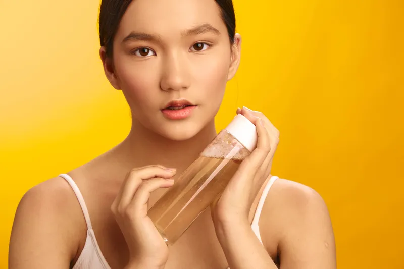 Asian woman stands against a vibrant backdrop, holding a clear skincare bottle near her face. She looks tranquil and confident, conveying beauty, wellness, and thoughtful care in a modern self care