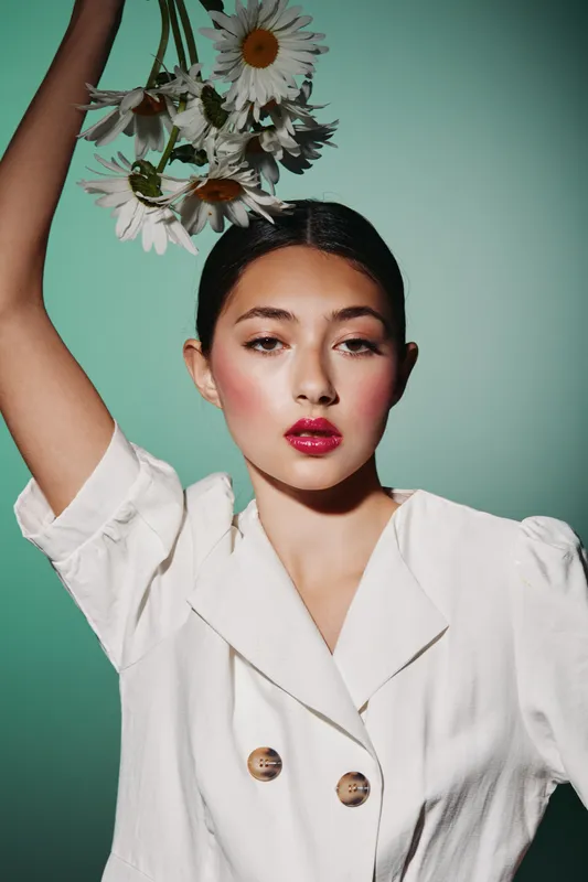 Fashionable young woman with red lips and rosy cheeks holds white daisies above her head. Portrait on green background in creative beauty and fashion concept studio.