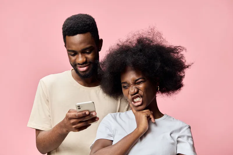Young African couple sharing a joyful moment while using a smartphone, showcasing emotions of happiness and surprise against a pink background