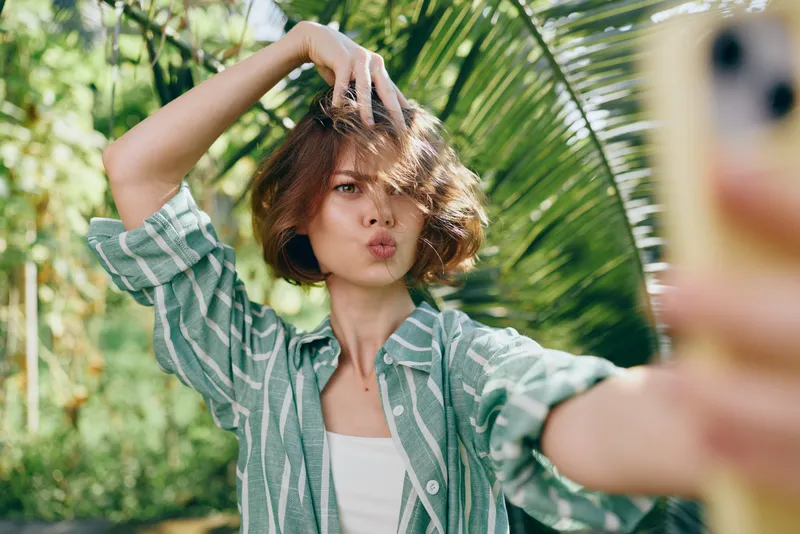 Woman wearing a striped green shirt takes a cheerful selfie outdoors, posing with a playful gesture and light smile, surrounded by lush greenery in a bright casual scene.