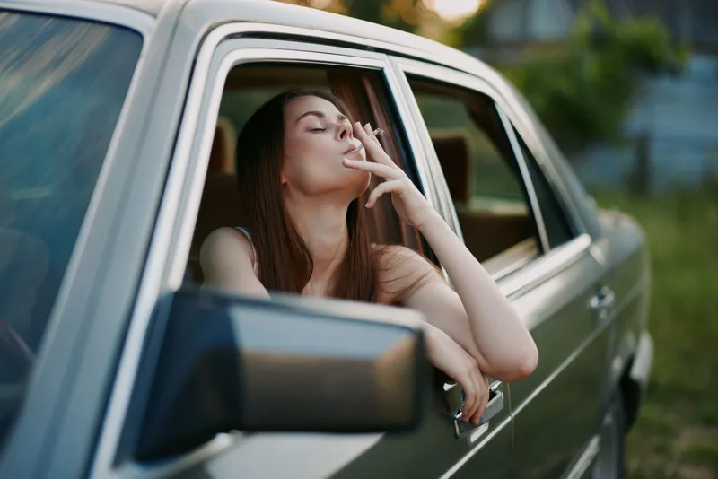 Young woman with long hair smoking a cigarette in a vintage car, evoking a sense of nostalgia and freedom in a serene outdoor setting.