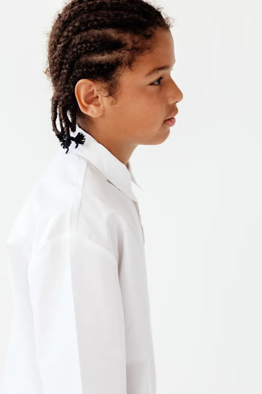 Модный маленький мальчик с заплетенными в косички волосами и в белой рубашке уверенно позирует на фоне гладкой белой стены