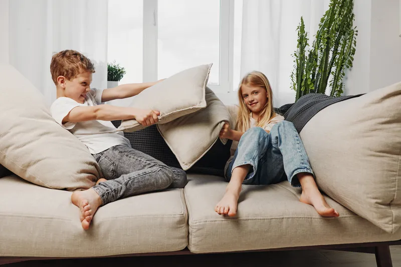 Two children having fun with a pillow fight on a beige sofa in a bright living room with plants and large windows in the background.