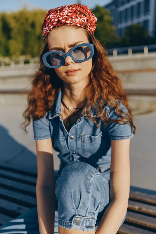 Lifestyle woman in modern boho-western denim street style wearing blue sunglasses and red patterned headband, captured with warm film color tones outdoors.