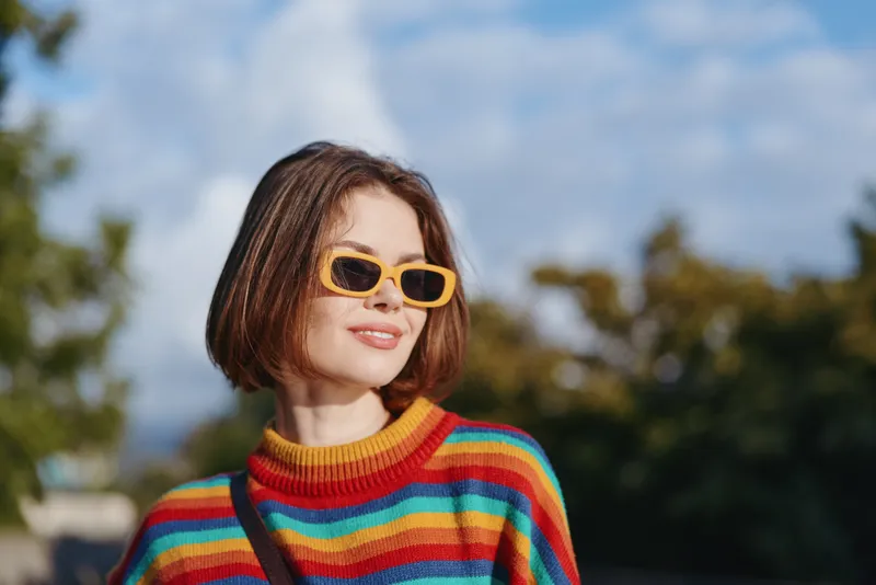 Woman in her mid twenties with short hair, smiling and wearing yellow sunglasses and a colorful striped sweater outdoors in a sunny park, casual lifestyle portrait for travel.