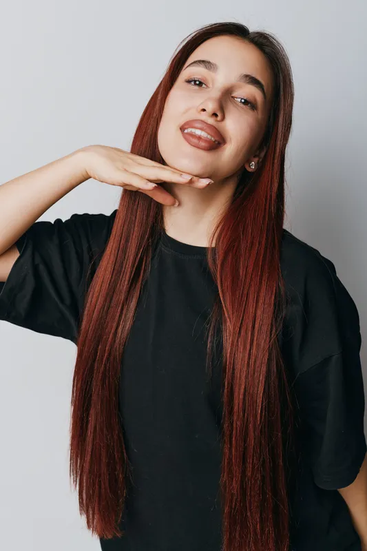 Young woman with long red hair posing with hand under chin and playful expression wearing black shirt on white background. Casual style and confident mood with natural makeup.