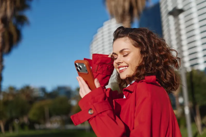 Young woman in a red coat smiles at her smartphone while capturing a cheerful moment outdoors in a bright city setting, radiating a candid, relaxed mood.