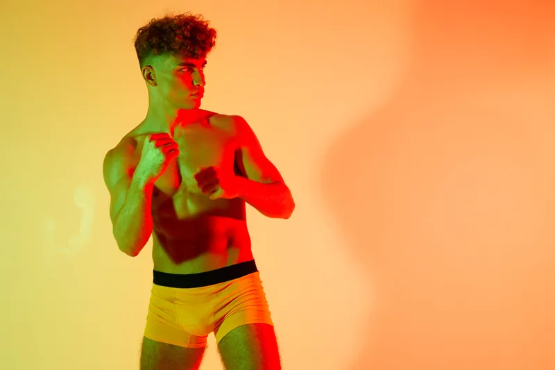 Young man with athletic body in yellow shorts posing for fitness training or boxing workout, colorful orange and red spotlight on background, muscle and strength concept.