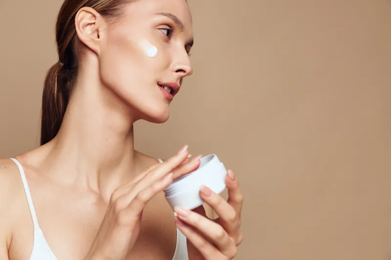 Woman applying eye cream with finger near cheek, holding white jar, close up on beige studio background, soft lighting, skincare and beauty routine, wellness and self care