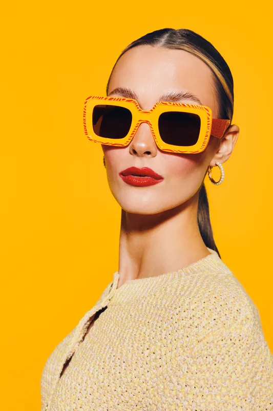 Stylish woman with slicked back hair wears oversized orange sunglasses and red lipstick. She poses confidently against a bright yellow background in a knitted beige top. Fashion concept.