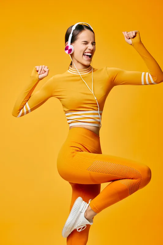 Energetic woman in bright orange activewear jumps mid-air against a vibrant orange backdrop, wearing headphones, celebrating fitness, sport, movement, and upbeat health.