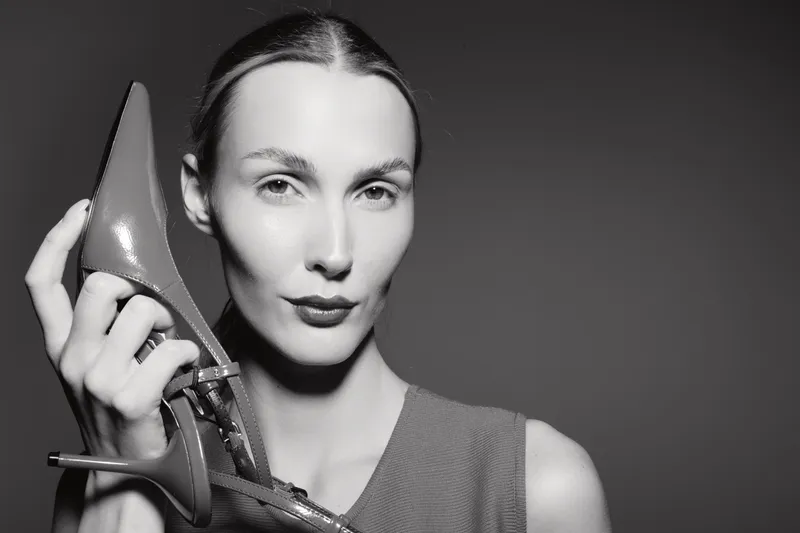 Portrait of white woman with light skin holding high heel shoe near face, studio lighting creates dramatic monochrome fashion concept.