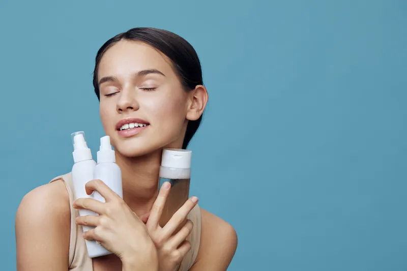 Young woman holding skincare products, showcasing healthy skin and a joyful expression against a blue background, emphasizing beauty and self care routines