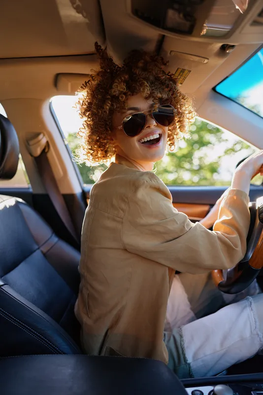 Cheerful woman in the car, driving with a wide smile, sunlit interior, retro film color, casual outfit, candid moment of joy and movement on a sunny day