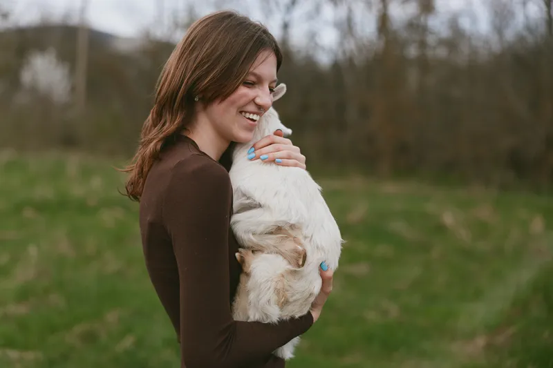 Cheerful woman outdoors, hugging a goat kid, warm smile, soft breeze, natural colors, grassy field backdrop, candid lifestyle moment, tender connection