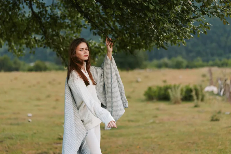 Woman lifestyle outdoor portrait in a field under a tree, wearing a cozy sweater and blanket, conveying relaxed fashion and serene mood in countryside scenery.