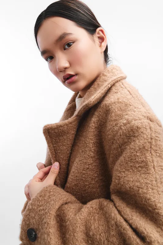 Asian woman wearing a warm camel coat stands against a clean background, presenting a poised and elegant look that conveys calm confidence in a studio portrait.