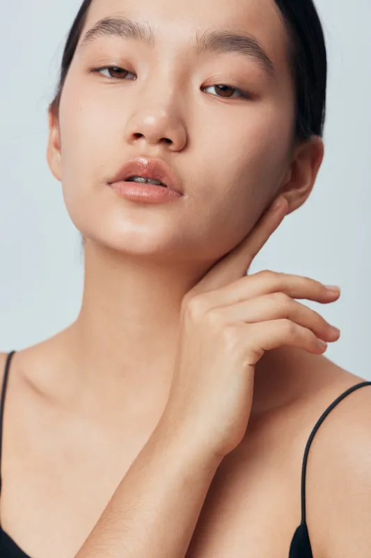 Portrait of young woman with natural skin and minimal makeup posing with hand near face on light background. Beauty model with clean complexion, smooth skin and serene expression in studio.