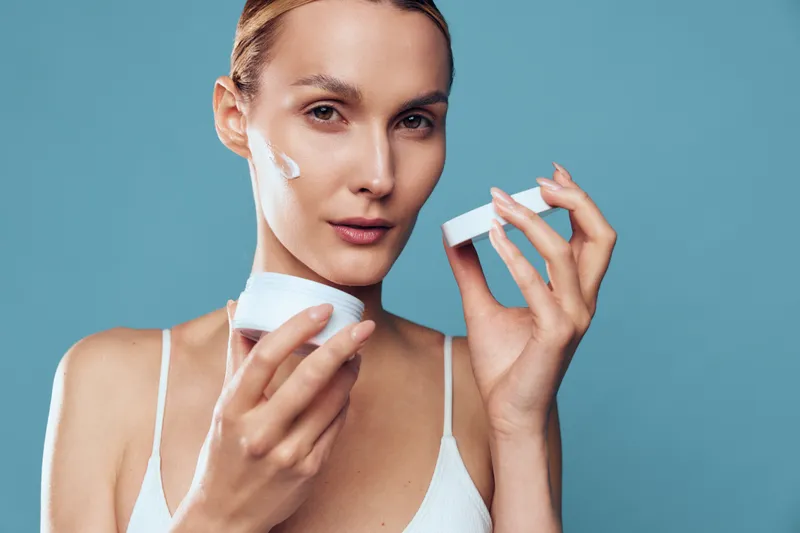 Young woman applying moisturizer cream on face, skincare routine, natural beauty, fresh skin, soft lighting, blue background, white top, focused expression, self care concept