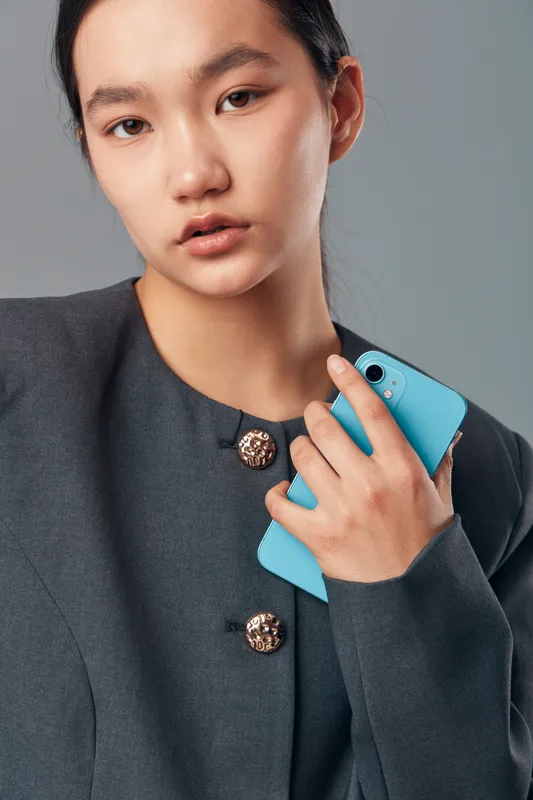Young woman with serious expression holding blue smartphone in hand wearing dark gray blazer with decorative buttons against neutral background in studio portrait.