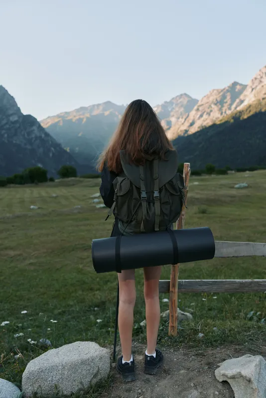 A lone hiker stands at a mountain trailhead with a backpack and rolled mat, prepared for exploration, gazing toward distant peaks under a calm sky.