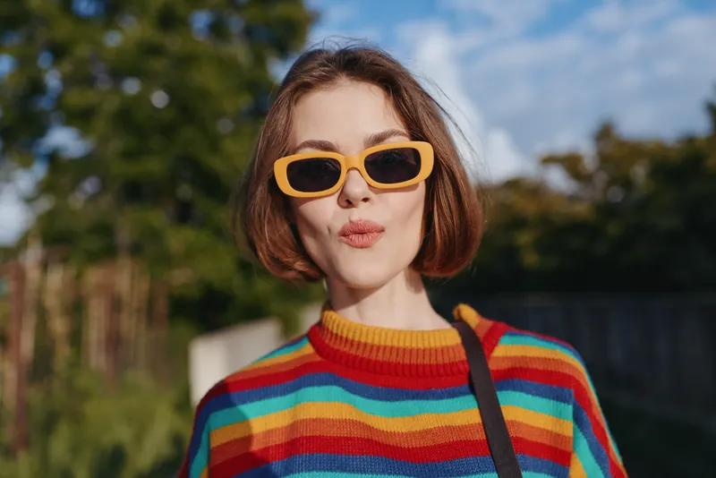 Woman in a colorful striped sweater and yellow sunglasses with a short bob haircut outdoors, playful puckered smile and casual shoulder bag, urban park setting with sunny lifestyle vibe.