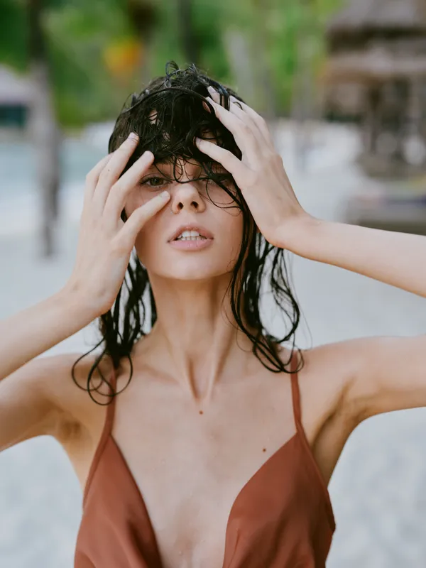 Wet woman with dark hair touching her face outdoors, wearing a brown swimsuit with a blurred green background and natural lighting.