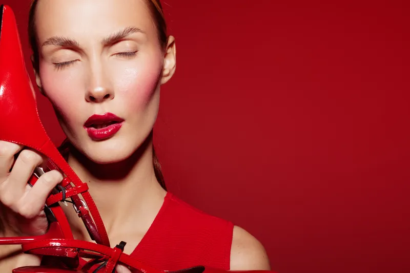 Woman with closed eyes holds red high heel shoes near face on red background. Concept of fashion, beauty, style, and elegance with vibrant color contrast and makeup.