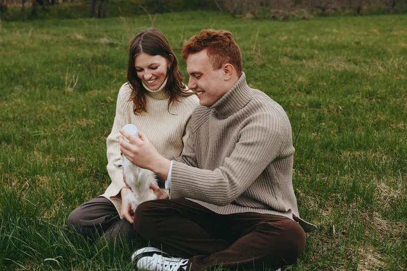 Young couple sitting on grass in cozy sweaters playing with a small white baby goat enjoying a relaxed lifestyle moment outdoors in a natural setting.