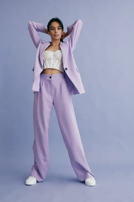 stylish woman wearing lilac suit with white corset and sneakers posing confidently against soft purple background in modern fashion editorial style