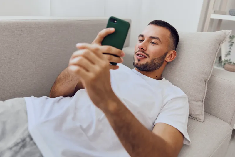 Relaxed man using smartphone while lying on a cozy sofa, showcasing modern lifestyle and technology in today's digital age. Perfect for tech and lifestyle themes.