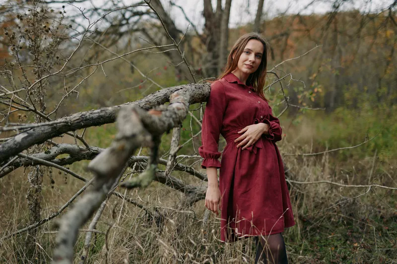 A natural outdoor portrait of a woman in a red dress leaning on a fallen tree, with a genuine hand on her belly and authentic expression, conveying honest human presence and calm mood.