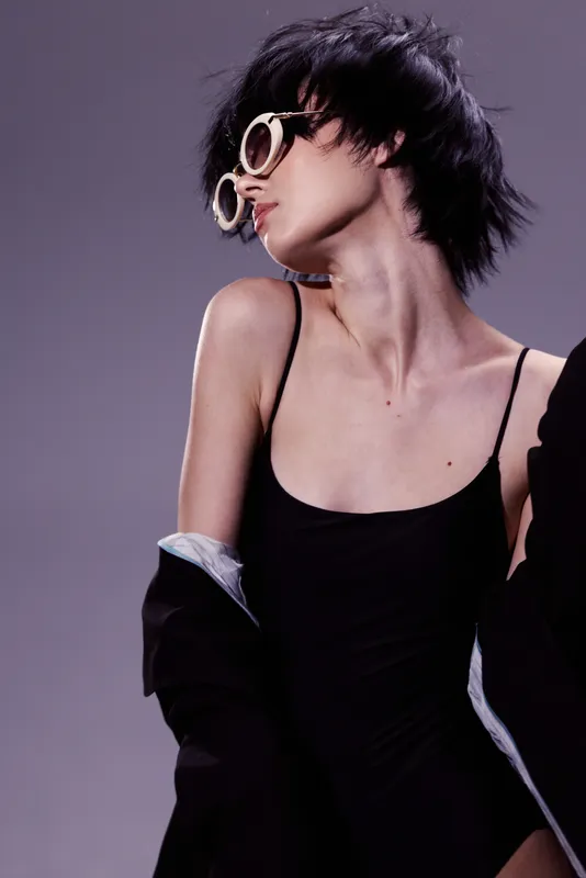 Stylish woman with short hair wearing sunglasses and black outfit posing against purple background