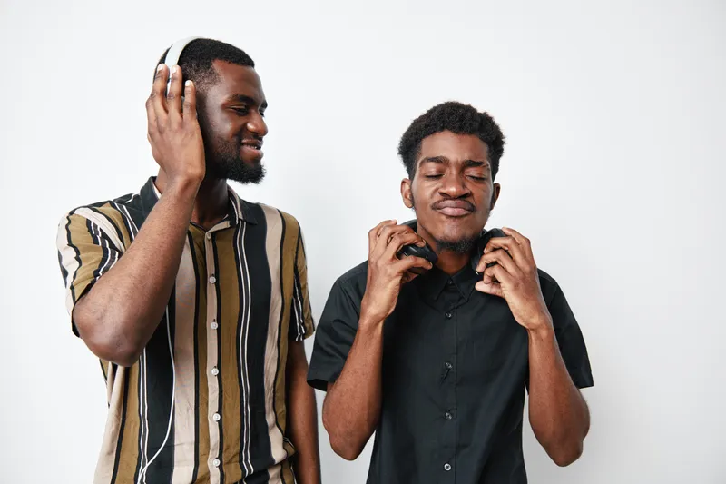 Two african men with dark skin enjoying music with headphones, smiling and relaxed in casual clothes against white background in studio. Friendship and leisure concept.