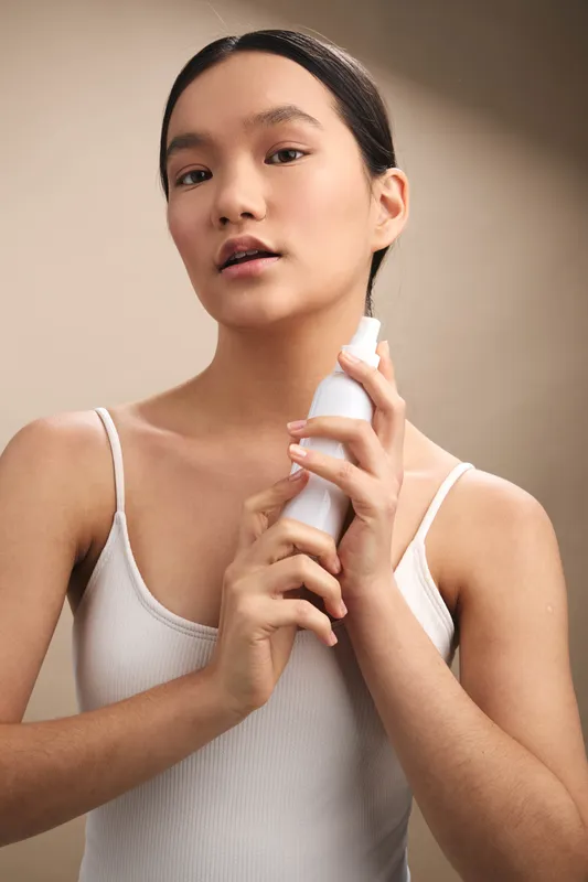 Asian woman in a simple tank top stands in a neutral studio, gently spraying a bottle to refresh her skin, conveying calm self care and fresh radiance.