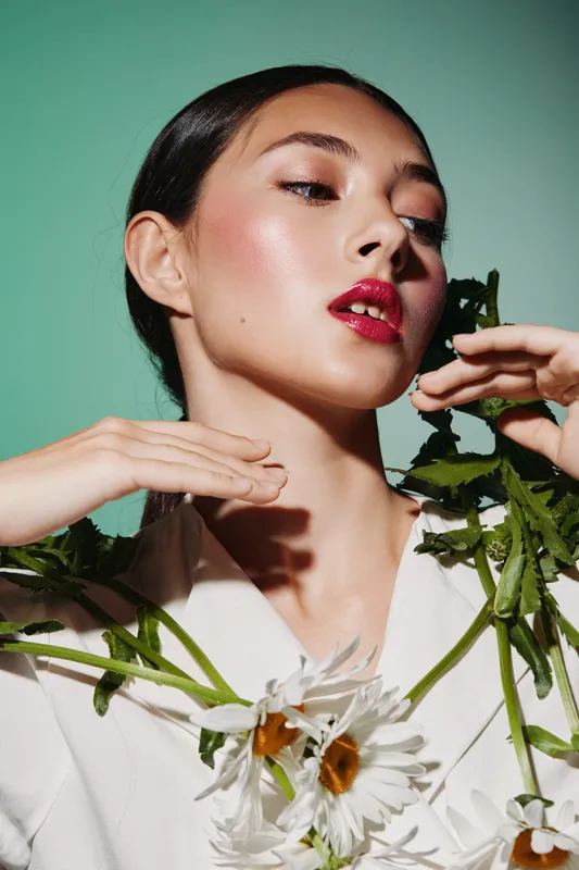 Young woman in white blazer with red glossy lips poses with white daisies on green background. Bold fashion beauty portrait with modern creative styling and soft shadows.