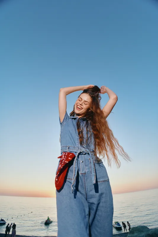 Lifestyle woman in modern boho-western denim street style smiling joyfully with long hair, red bandana, and serene ocean background in warm film color tones.