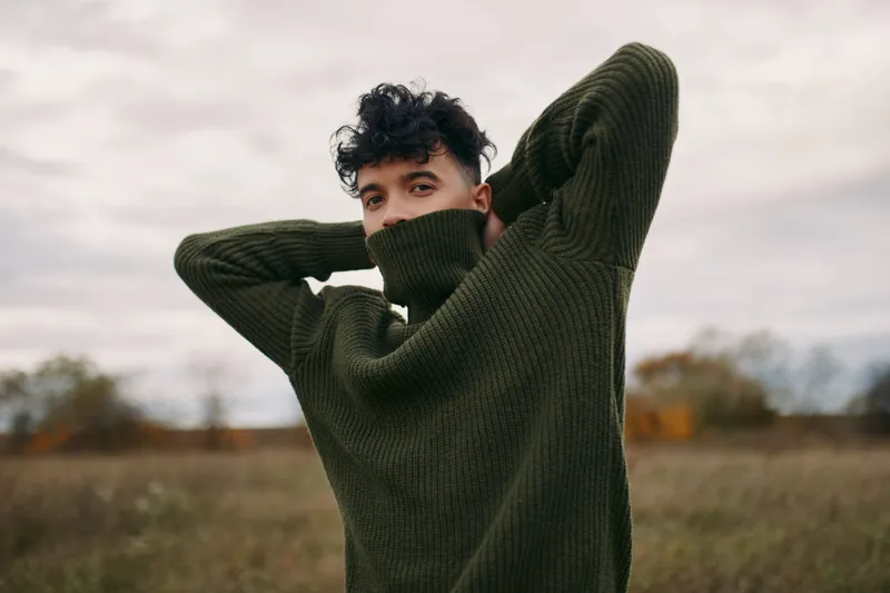[Portrait of a young man in a green knit sweater standing in an open field, captured with natural light to convey authenticity and credibility through honest expression and texture.]