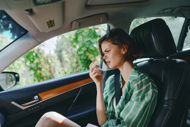 A woman sits in a car seat, talking on a phone with a thoughtful, anxious expression as soft light filters through the window.