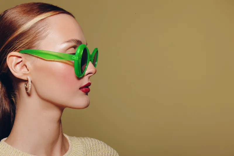 Profile of a stylish woman wearing bright green sunglasses and gold hoop earrings against a plain beige background. Concept of fashion, accessories, and modern style.