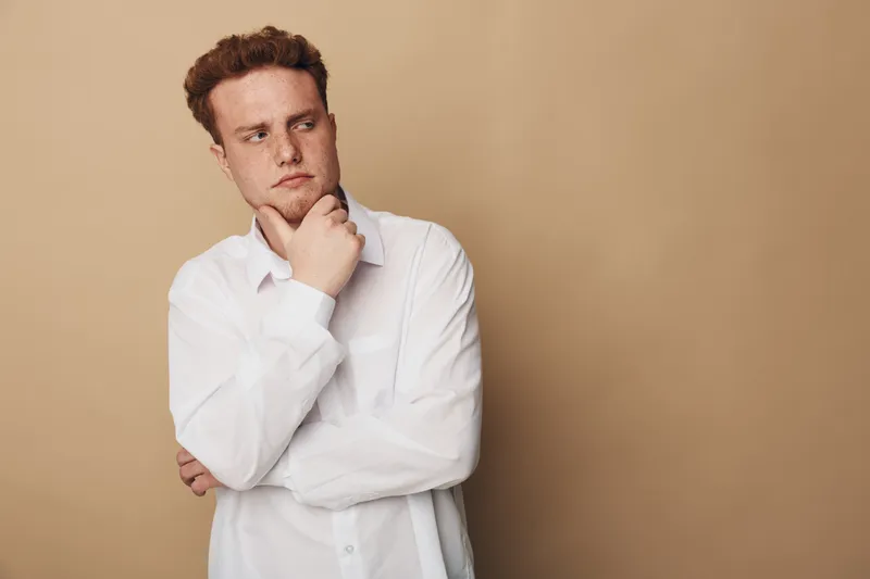 Thoughtful young man with red hair wearing white shirt looks away with hand on chin. Neutral beige background. Concept of contemplation, decision making, and introspection.