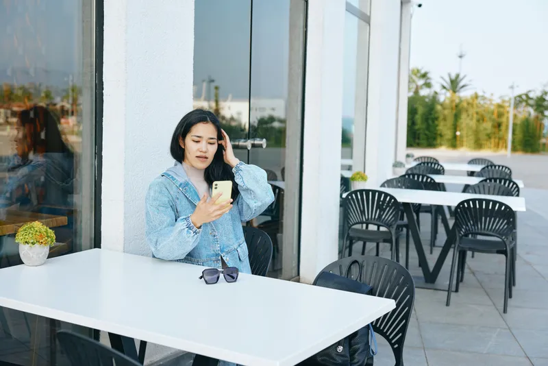 Woman sits at a bright outdoor cafe, checking smartphone, wearing blue patterned shirt. Sunglasses rest on white table, reflective windows behind, calm and casual mood, daytime urban setting, relaxed