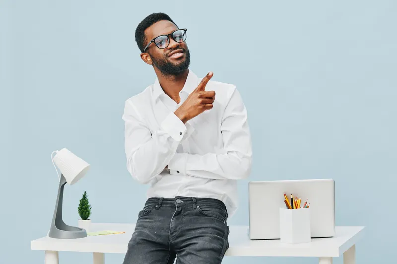 Professional African American man with glasses wearing white shirt, leaning on desk, demonstrating thoughtful expression and pointing finger, in modern office interior with laptop and workspace equipm